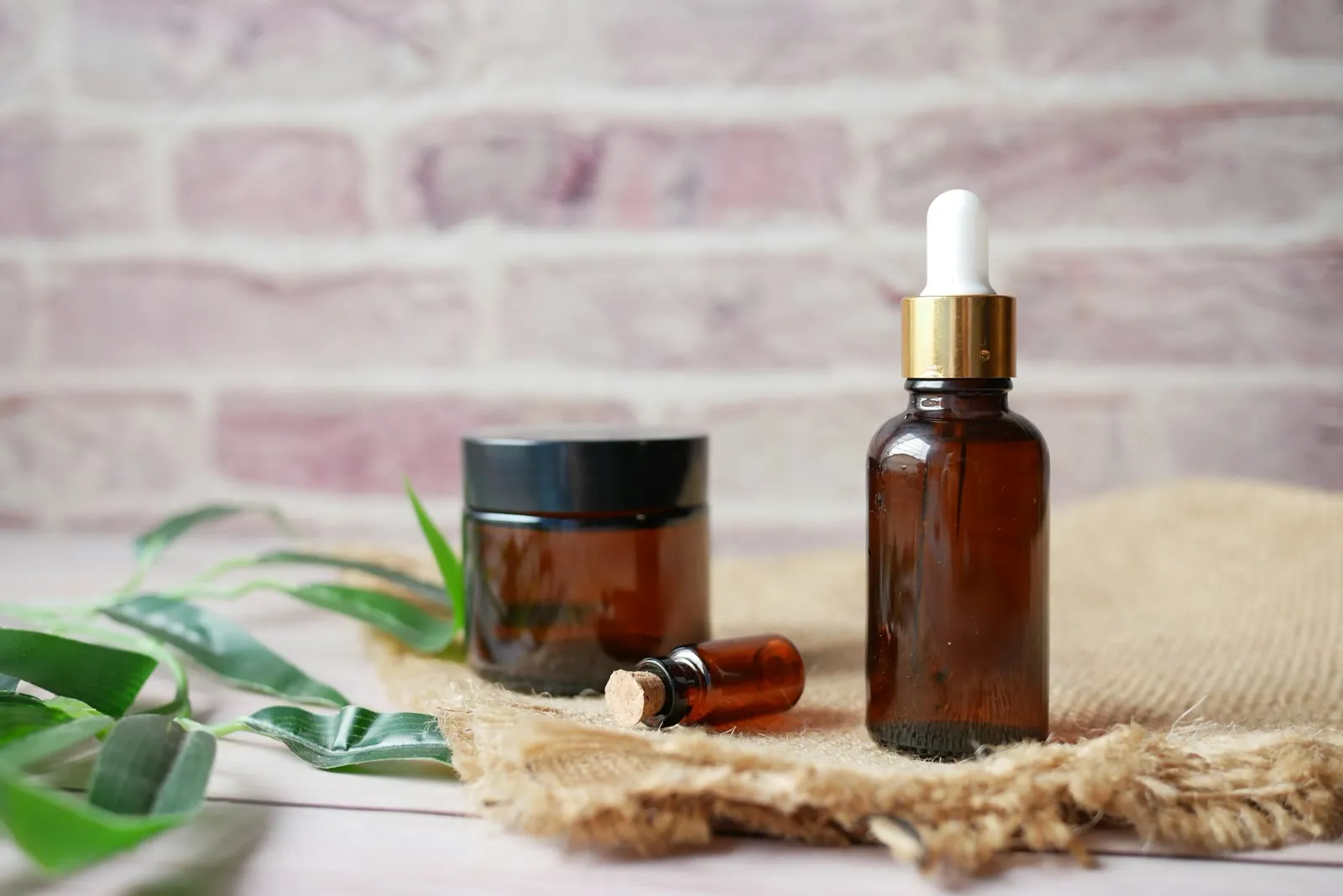 Amber glass serum bottles and a jar on a linen cloth beside green leaves