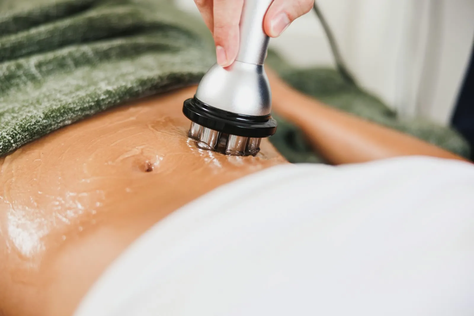 A handheld treatment device moving over gel on a client’s abdomen, with a green towel below