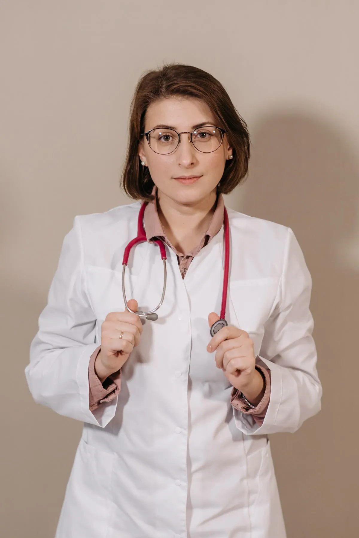 A physician in a white coat and glasses holding a stethoscope, against a warm beige wall