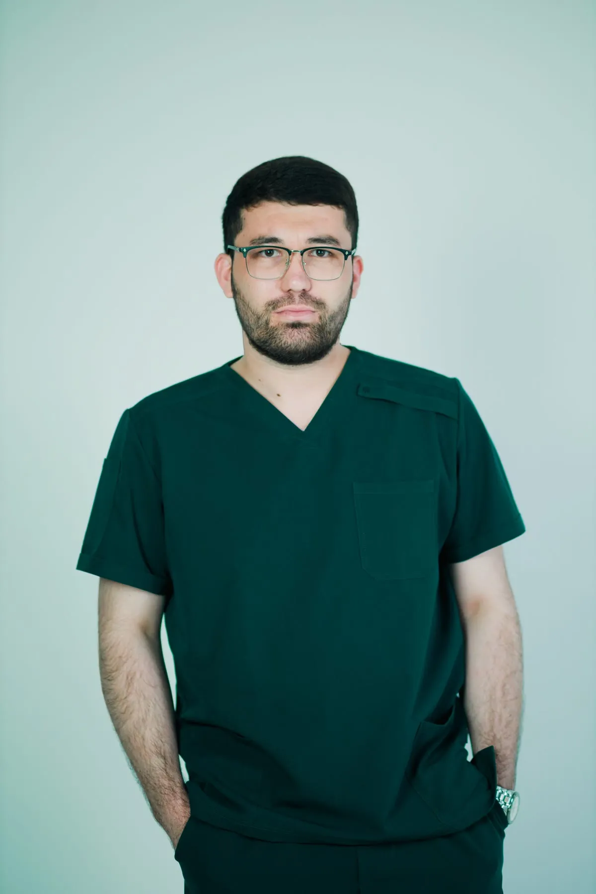 A clinician in dark green scrubs and glasses, standing with his hands in his pockets